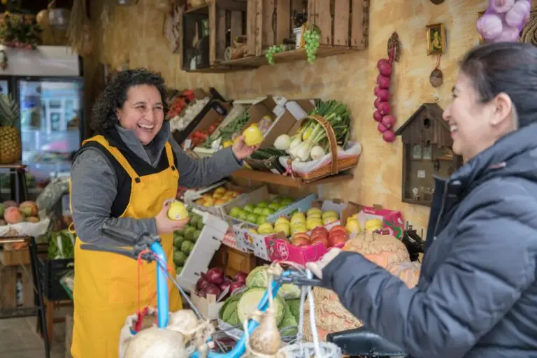 Two women smiling and exchanging fresh produce at a vibrant greengrocer's market.